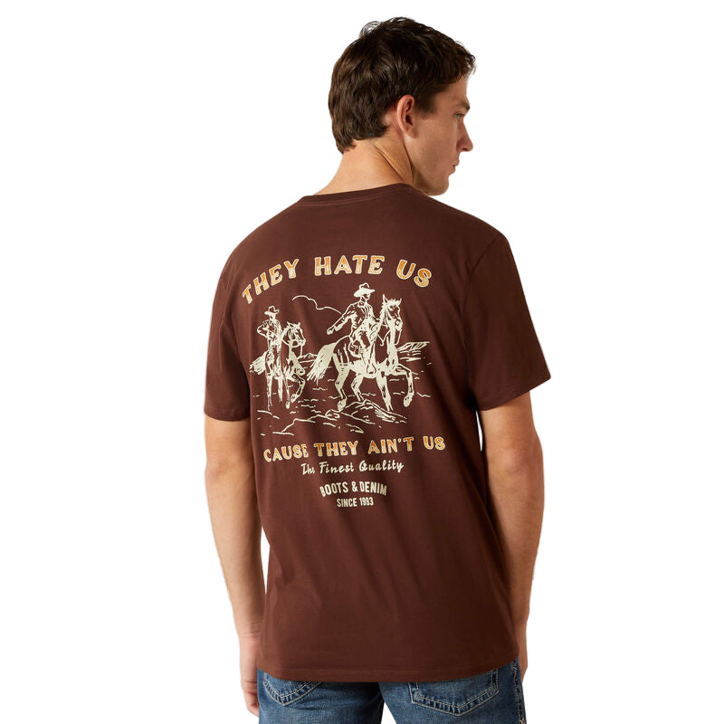 Man wearing a brown t-shirt with graphic design and text on the back, standing against a beige background.