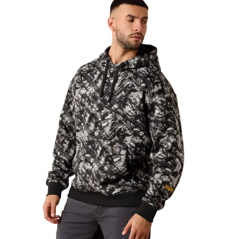 A man wearing a black and white abstract patterned hoodie with black ribbed cuffs, collar, and hem, paired with dark gray pants, photographed against a solid light beige background.