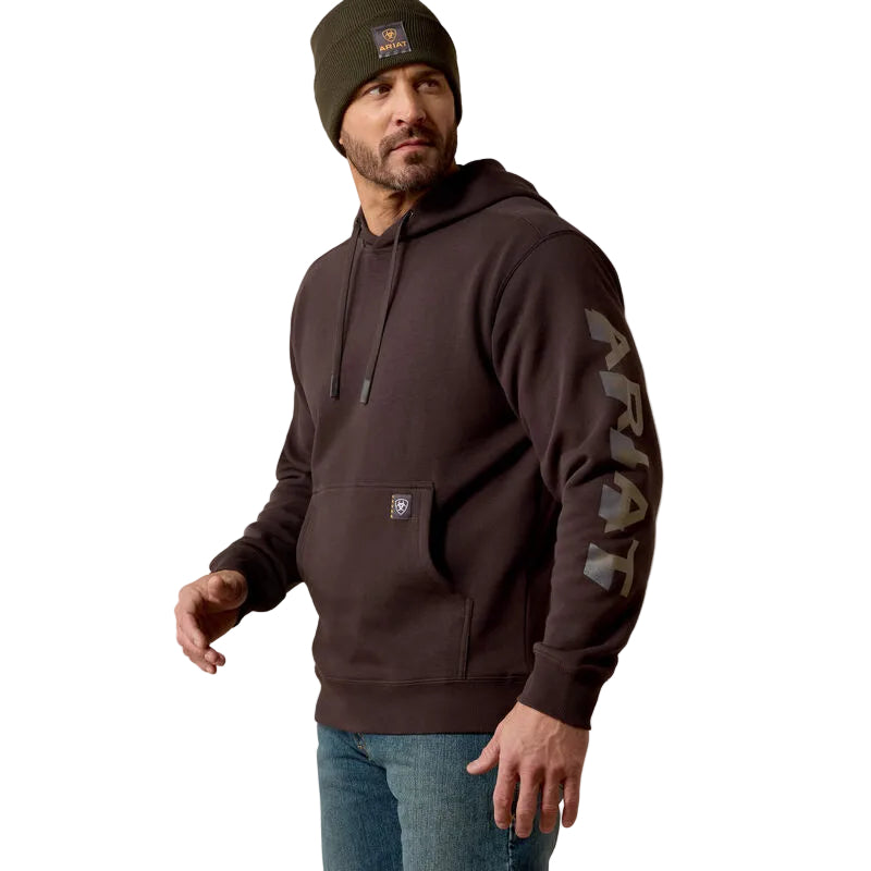 A man wearing a dark brown heavy-duty hoodie with a large gray 'ARIAT' graphic on the left sleeve, paired with blue jeans and an olive green beanie. The hoodie features a kangaroo pocket and a small brand patch on the pocket.