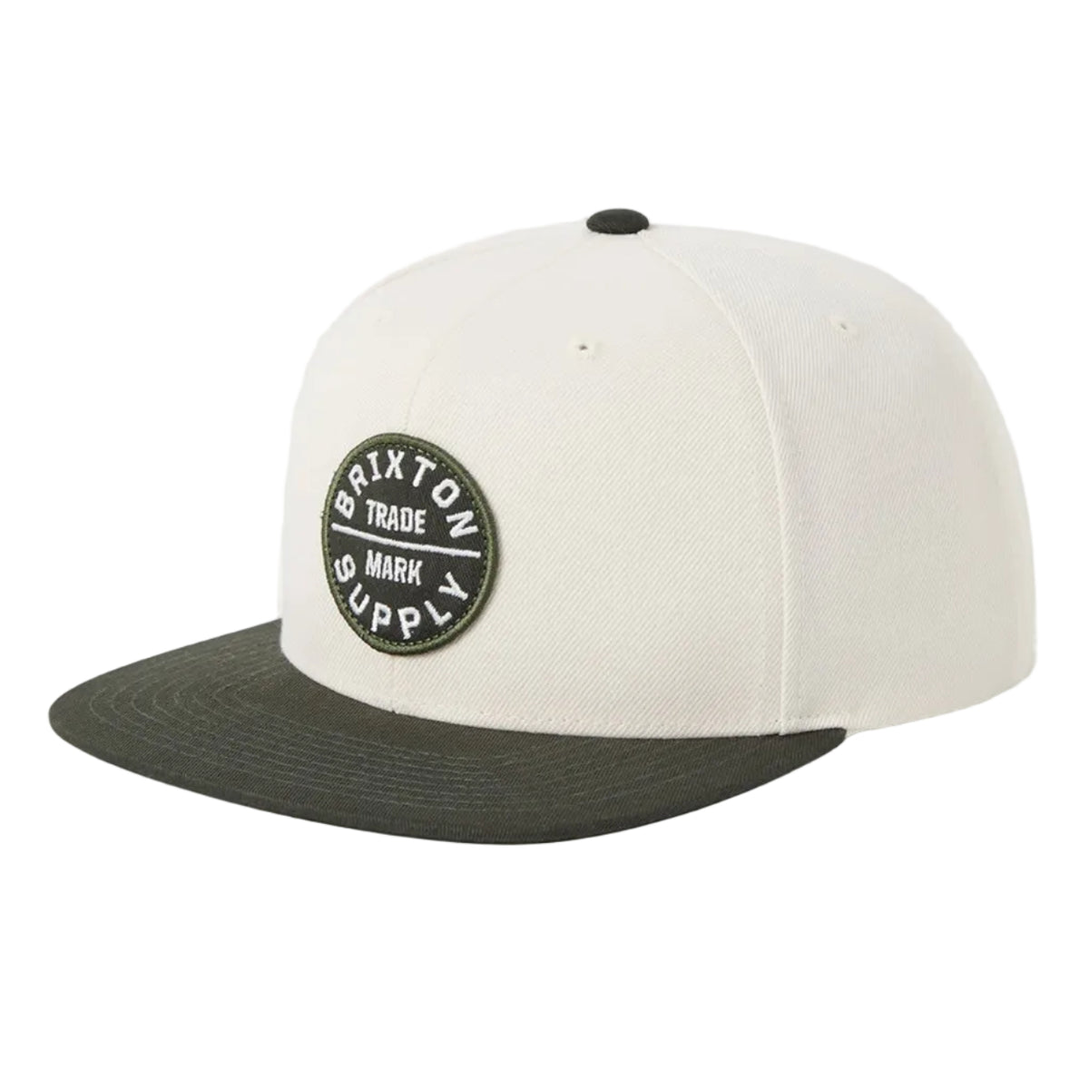 A structured baseball cap with an off-white crown and a dark olive green flat brim, featuring a circular black and white embroidered patch on the front that reads 'Brixton Made Supply'.