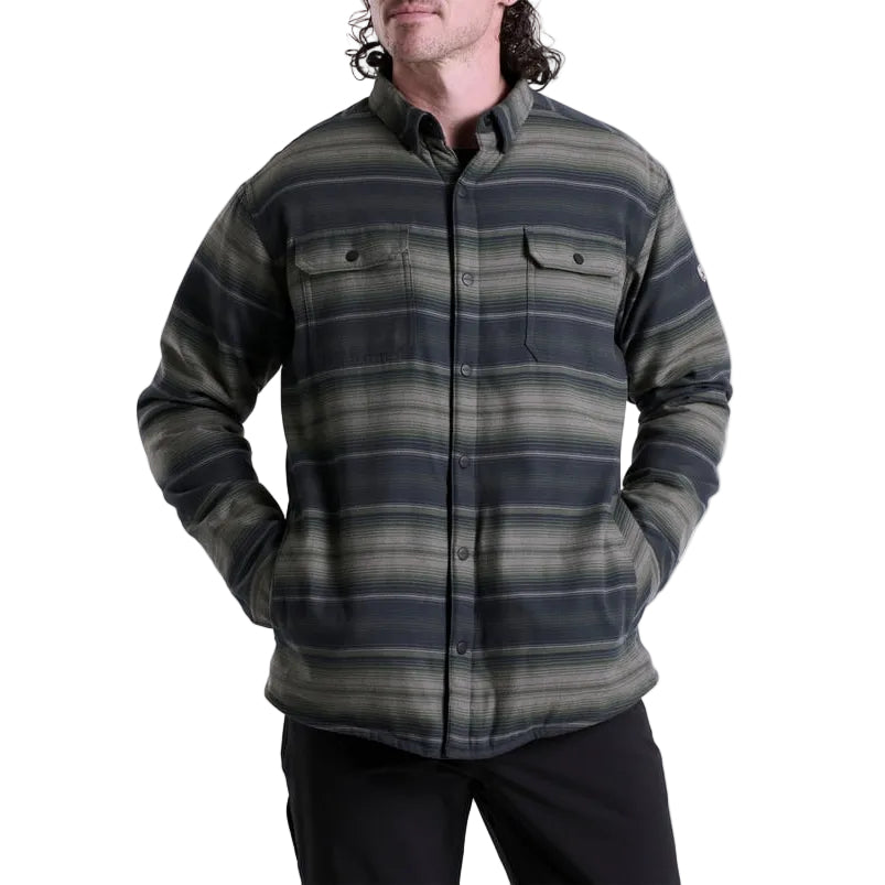 A man wearing a long-sleeve button-up flannel shirt with horizontal stripes in dark green, gray, and black, paired with black pants, against a plain white background.